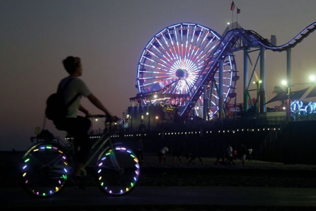 A cyclist rides along a path with lights on the wheels on Santa Monica Pier with Ferris wheel in multiple lights to illustrate NIH looks to slim down peer review in bid for equity