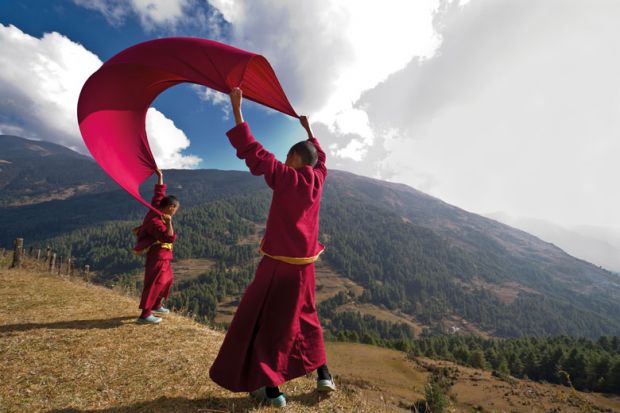Young monks wearing red clothes play as if flying a kite with a blanket on a mountain slope to illustrate Nepalese peak a new red flag for Australian enrolment