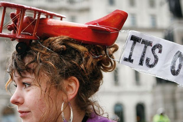 A protestor wears a toy aeroplane on her head with a banner reading  'It's over' to illustrate  ‘Set institutional carbon budgets to cut flying and support rail travel’
