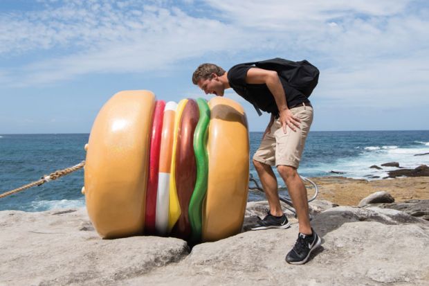 A person  mocks taking a bite out of "What a Tasty Looking Burger" by James Dive at Sculpture By The Sea in Sydney, Australia to illustrate Campus catering ‘too expensive and unhealthy’