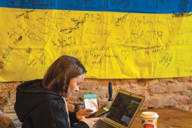 A woman works on a laptop in a room with a  Ukrainian flag on the wall to illustrate International help ‘must not kill Ukrainian system with kindness’
