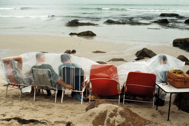 People on deckchairs shelter under plastic sheets on beach
