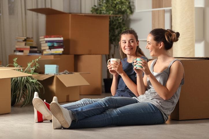 two female students sitting on the floor with their moving boxes, smiling and drinking from mugs