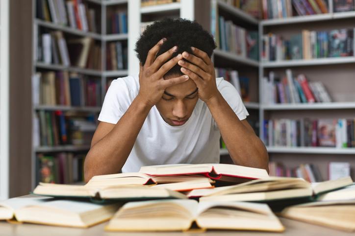 young man with his head in his hands reading lots of books in the library